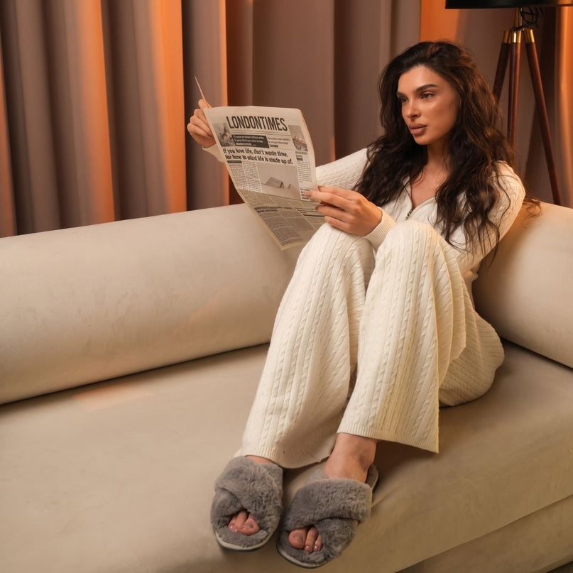 Woman reading a newspaper on a cream sofa wearing BELSTA The Ash Fluffy Slippers, grey plush open-toe slides