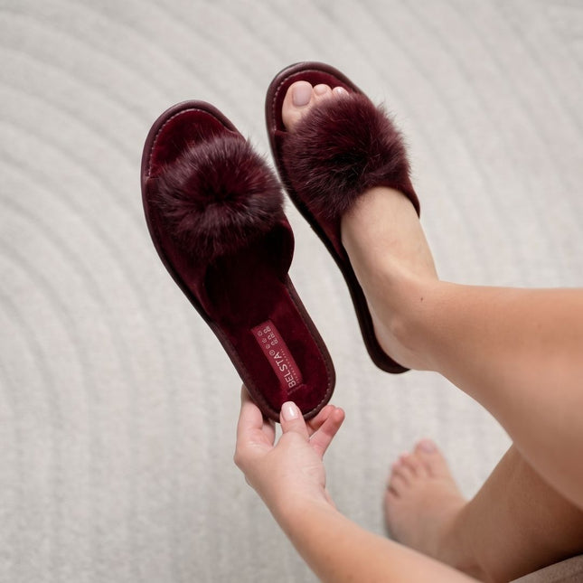 Woman putting on BELSTA The Pom-Pom Slippers — burgundy velour slides with plush pom-poms, lifestyle shot