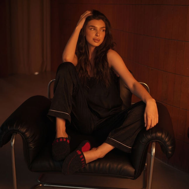Woman in black pajamas in a leather armchair wearing BELSTA The Ember Felt Slippers, charcoal felt slippers with red trim